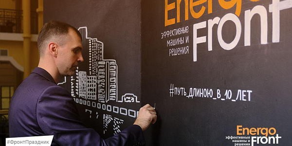 Мужчина в костюме пишет мелом хештег на чёрной стене стенда Energo Front